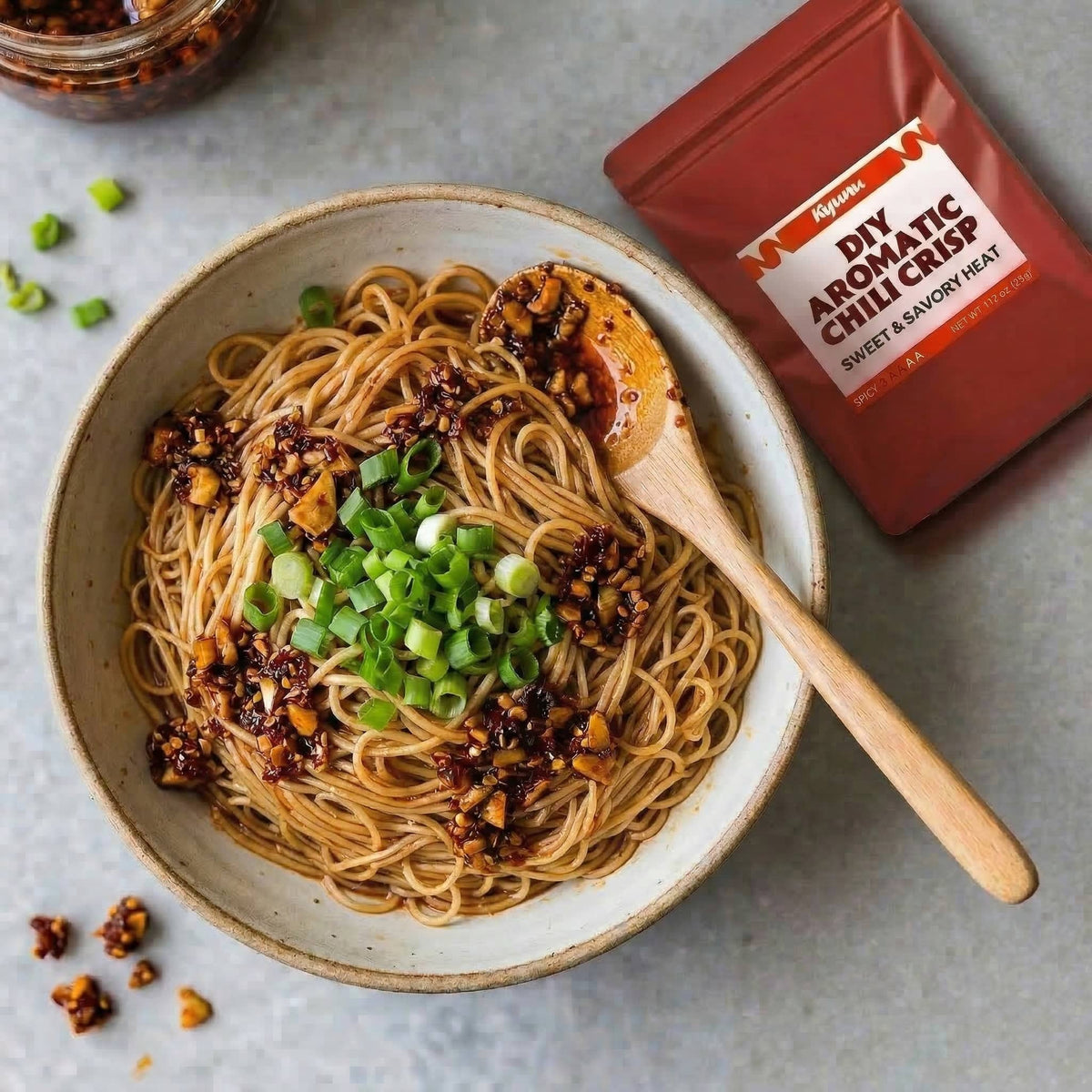 Bowl of noodles tossed with chili crisp and scallions beside the Kyunu DIY Aromatic Chili Crisp pouch