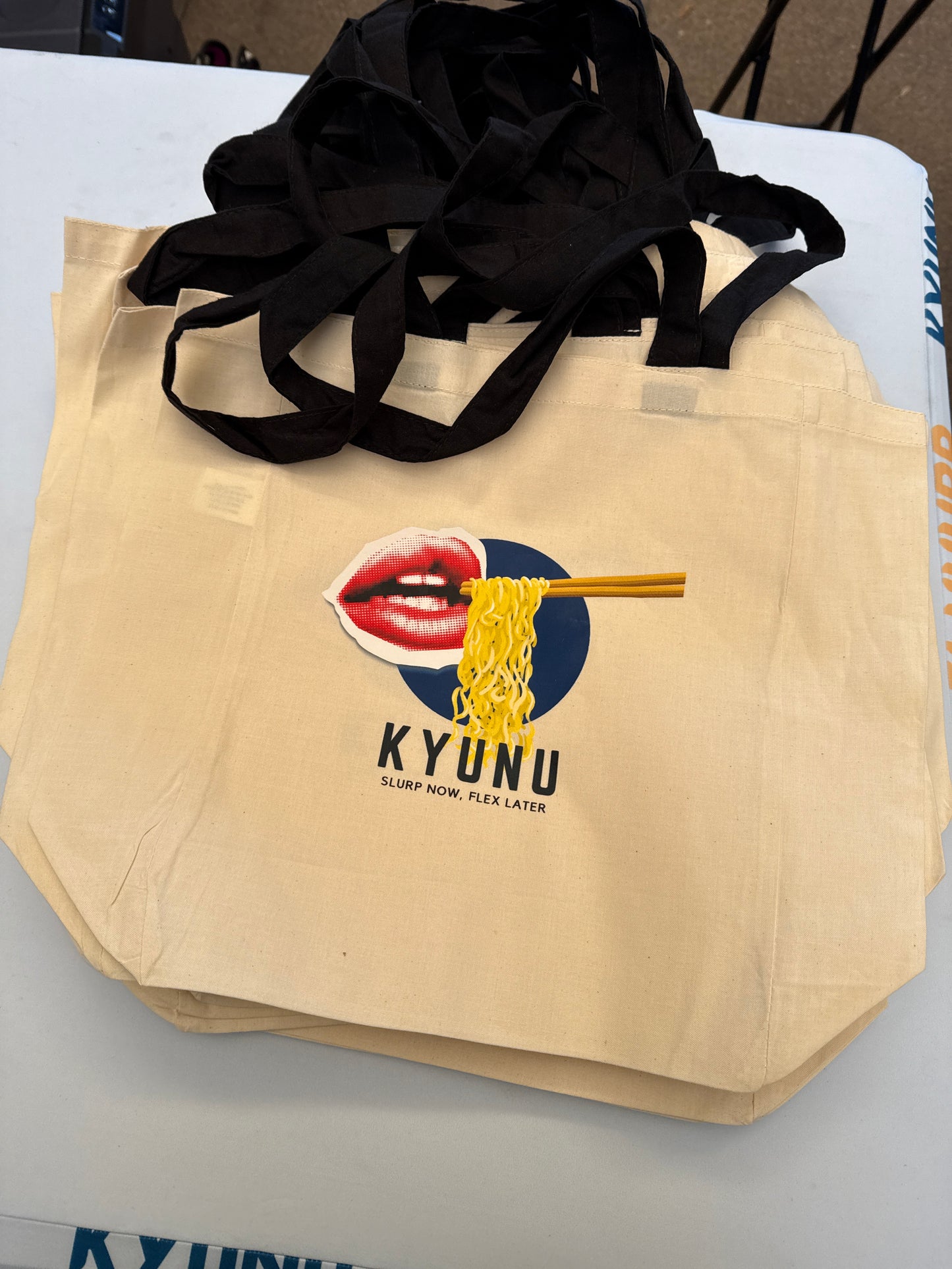 Stack of natural canvas Kyunu tote bags printed with the lips and noodles logo and Slurp Now, Flex Later