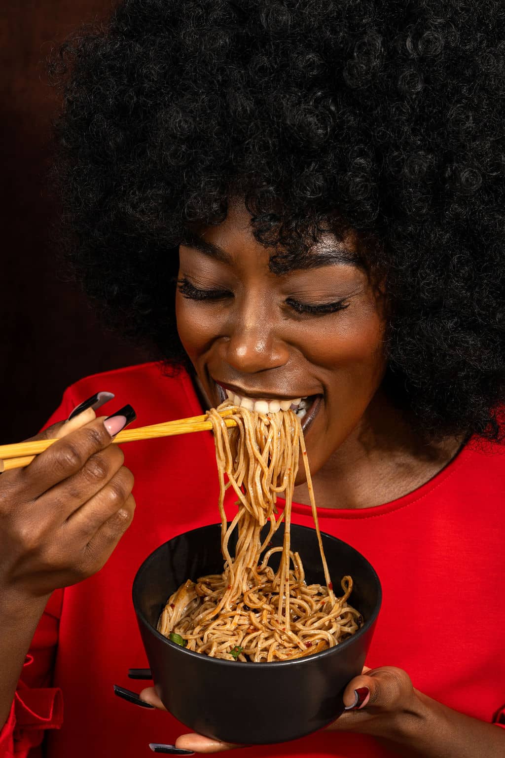 Woman eating noodles with chopsticks from a black bowl against a dark background